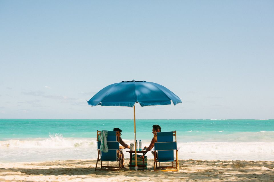 Two guests seated beneath a blue Beach Day umbrella facing the ocean